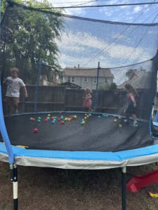 Kids are playing on a netted-in trampoline with colorful ball pit balls around their feet.