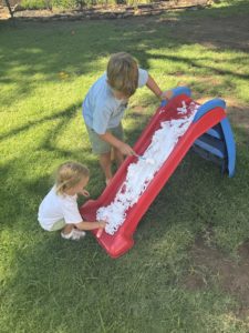 A brother and sister are playing outside and painting their slide with shaving cream. Ideas to beat the heat with little kids. 
