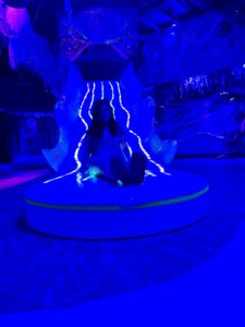 A teenager at a bottom of a neon blue lit slide at Factory Obscura.